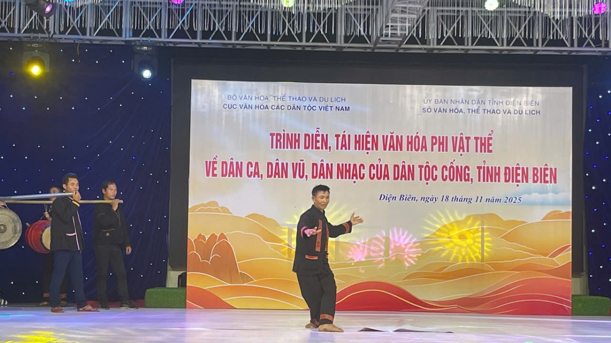 Một nghệ nhân nam dân tộc Cống đang biểu diễn trên sân khấu với động tác múa mạnh mẽ, thể hiện tinh thần của dân vũ truyền thống. Phía sau là hình ảnh phông nền rực rỡ với dòng chữ thể hiện nội dung sự kiện "Trình diễn, tái hiện văn hóa phi vật thể về dân ca, dân vũ, dân nhạc của dân tộc Cống, tỉnh Điện Biên". Nguồn: Ảnh do đơn vị tổ chức cung cấp.